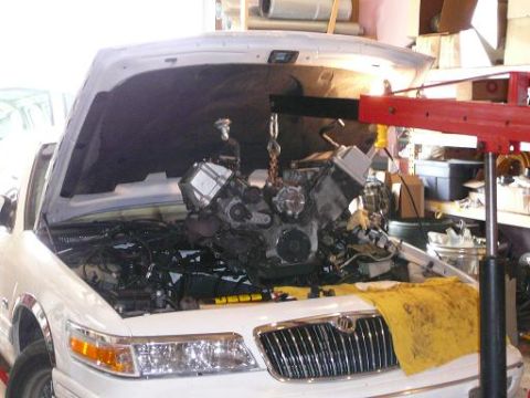 1997 Mercury Grand Marquis - White Mercury sedan in a garage with its V engine lifted out on an engine hoist above the empty engine bay.