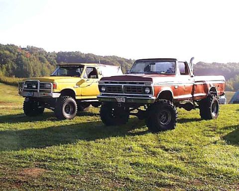 1988 Ford Bronco - Two lifted trucks, one yellow and one red, sit on a grassy field with trees in the background.