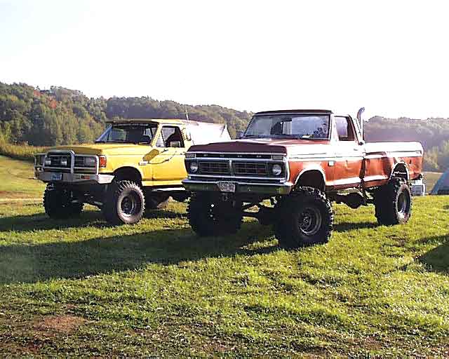 Yellow lifted Ford Bronco parked beside an orange 1970s Ford 4x4 pickup on a grassy hill