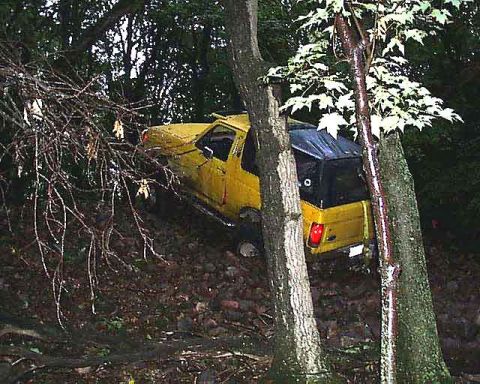 1988 Ford Bronco - A yellow SUV is partially off the road and stuck on a steep, wooded incline.