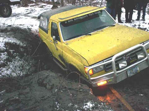 1988 Ford Bronco - A yellow Ford Bronco is stuck in a muddy ditch, with people watching in the background.