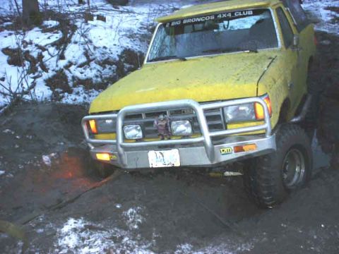 1988 Ford Bronco - A yellow 1988 Ford Bronco is stuck in the mud, with a "BIG BRONCOS 4X4 CLUB" sticker visible on its windshield.