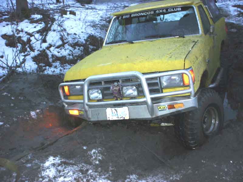 Yellow 1988 Ford Bronco stuck in mud with "BIG BRONCOS 4X4 CLUB" sticker on windshield