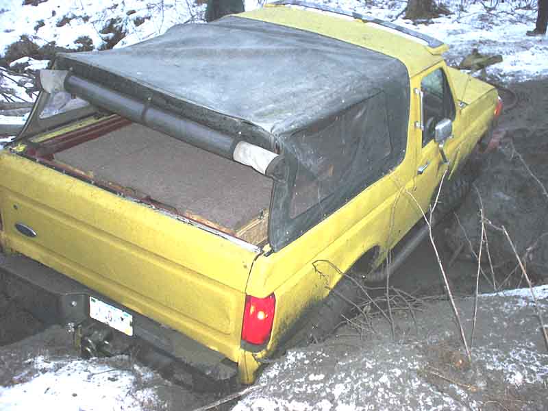 Yellow Ford Bronco with soft top folded back, stuck to the frame in a snowy rut