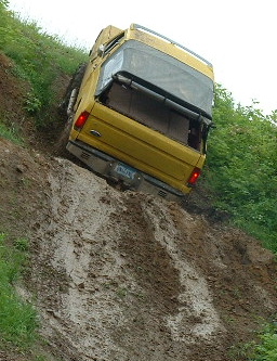 1988 Ford Bronco - A yellow SUV is stuck on its side on a steep, muddy incline, with trees and grass visible on the banks.