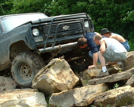 1988 Ford Bronco - A group of people examine the front of a black off-road truck that appears to be stuck on a rocky terrain.