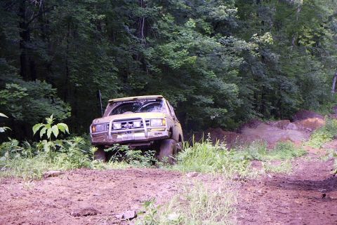 1988 Ford Bronco - A yellow SUV is stuck in the mud on a wooded trail, its front end partially obscured by vegetation.