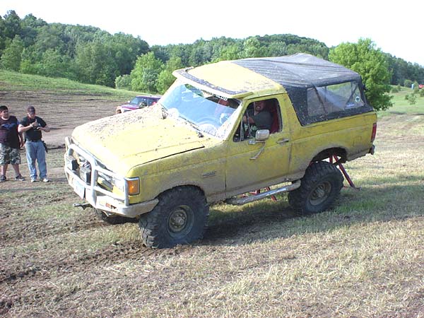 Muddy yellow Ford Bronco with a soft top, lifted suspension, and off-road tires, stuck in mud. Two people stand nearby. Another red SUV is visible in the background