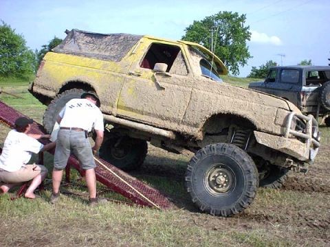 1988 Ford Bronco - Two people assist a heavily mud-covered off-road vehicle on ramps, with another vehicle in the background.
