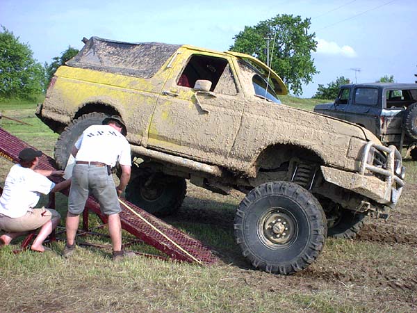 Muddy yellow off-road vehicle on ramps with two people nearby. Another dark vehicle is parked behind it