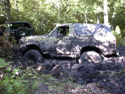 1988 Ford Bronco - Two off-road vehicles are stuck in thick mud on a wooded trail, with one vehicle almost completely submerged.