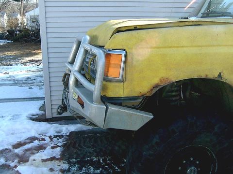 1988 Ford Bronco - A yellow truck with a brush guard and large tires is parked next to a building in the snow.