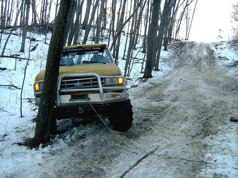 1988 Ford Bronco - A yellow Ford Bronco is winched to a tree while navigating a snowy, wooded incline.