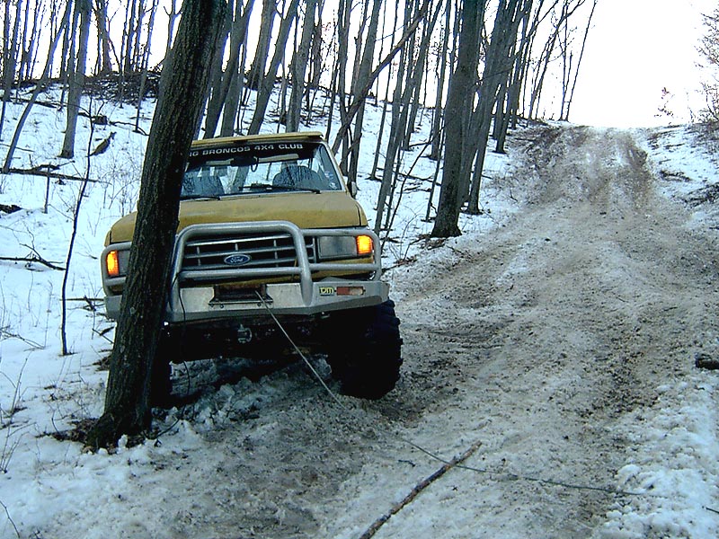 Yellow Ford Bronco with winch on snowy hill, tree in foreground. Decal on windshield says '1980 Broncos 4x4 Club'
