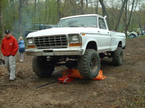 1988 Ford Bronco - A lifted white pickup truck sits on a jack in an outdoor setting with trees and tents in the background.