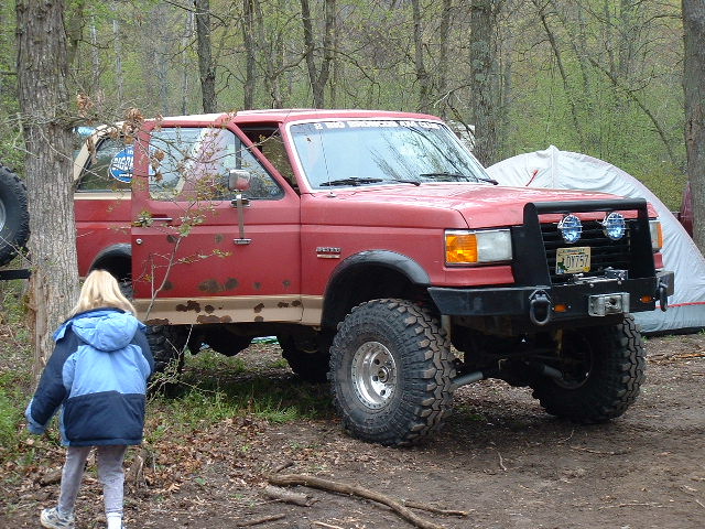 Lifted red Ford Bronco with winch bumper and mud tires parked in the woods, front three-quarter