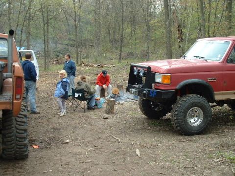 1988 Ford Bronco - A red 1988 Ford Bronco is parked in a wooded area with people gathered around a campfire.