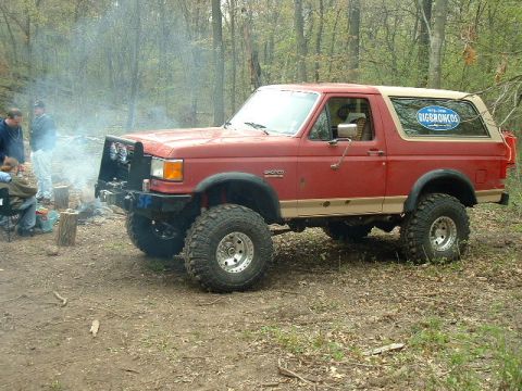 1988 Ford Bronco - A red Ford Bronco with a tan top and off-road tires sits in a wooded area next to a campfire.