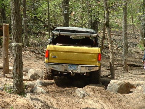 1988 Ford Bronco - This yellow SUV is shown from the rear, driving over a rough, wooded trail with large rocks.