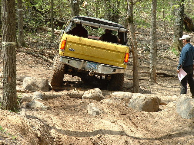 Lifted yellow Ford Bronco flexing over a rock ledge on a wooded trail, rear view