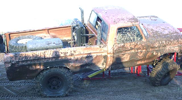 Mud-covered lifted pickup with exhaust stack climbing a red steel ramp obstacle