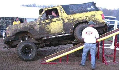 1988 Ford Bronco - A heavily muddied yellow SUV is positioned on a ramp, suggesting an off-road event.