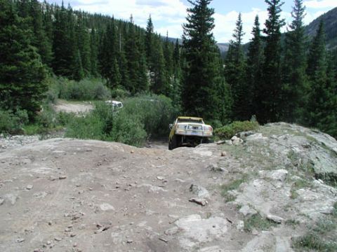 1988 Ford Bronco - A yellow pickup truck drives on a challenging, rocky mountain path with dense pine forest surrounding it.