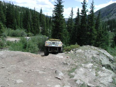 1988 Ford Bronco - An off-road vehicle navigates a challenging rocky path in a mountainous, forested area.