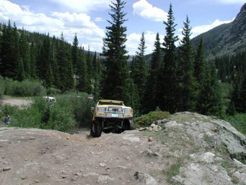 1988 Ford Bronco - A yellow off-road vehicle navigates a rocky mountain trail surrounded by pine trees.