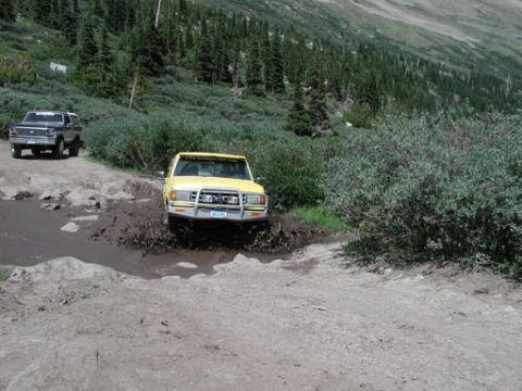 1988 Ford Bronco - Two trucks navigate a muddy trail with dense trees and a mountain slope in the background.