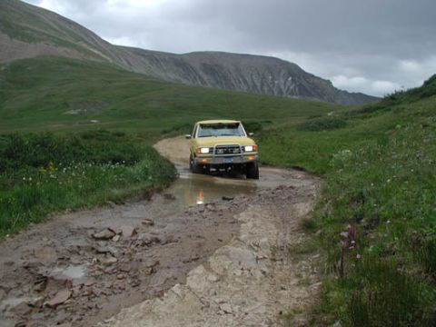 1988 Ford Bronco - A yellow pickup truck navigates a wet, rocky path in a mountainous landscape under a cloudy sky.