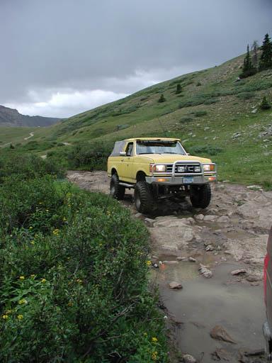 1988 Ford Bronco - A yellow 1988 Ford Bronco navigates a challenging, rocky mountain path under a cloudy sky.