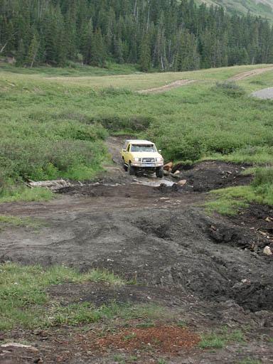 1988 Ford Bronco - A yellow SUV drives through a challenging off-road terrain with dense forest ahead.