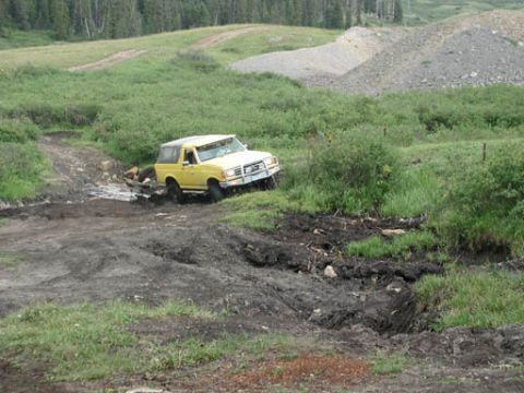 1988 Ford Bronco - This yellow SUV is driving through a muddy, off-road trail with vegetation and hills surrounding it.