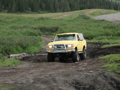 1988 Ford Bronco - A yellow 1988 Ford Bronco navigates a muddy trail surrounded by greenery.