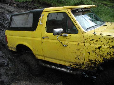 1988 Ford Bronco - Mud splatters cover a yellow 1987 Ford Bronco with a black soft top as it is stuck in thick mud.