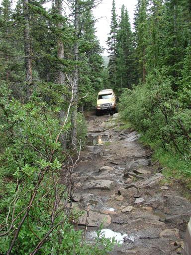 1988 Ford Bronco - A yellow SUV carefully drives up a challenging, muddy, and rocky path surrounded by dense trees.