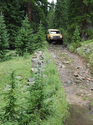 1988 Ford Bronco - A yellow SUV navigates a rough, wooded trail, surrounded by evergreen trees.