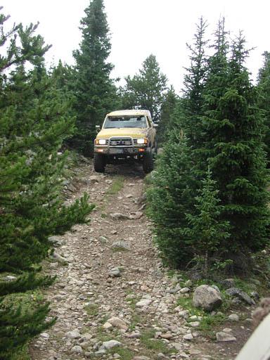 1988 Ford Bronco - A yellow 1988 Ford Bronco navigates a rocky, tree-lined dirt road.