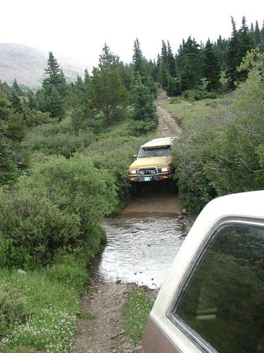 1988 Ford Bronco - Two vehicles navigate a mountain trail, crossing a small stream amidst dense forestation.