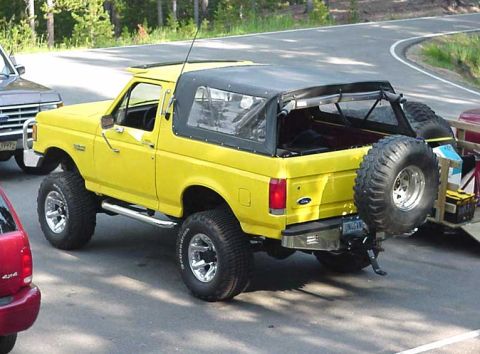 1988 Ford Bronco - A lifted yellow Ford Bronco with a black soft top is parked on a road next to other vehicles.