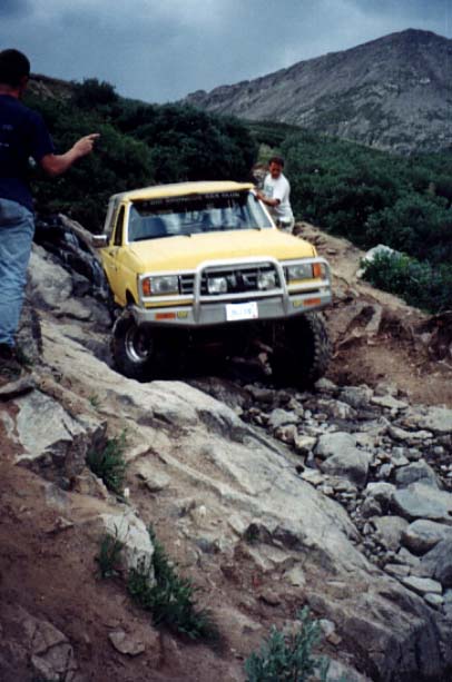 1988 Ford Bronco - A yellow pickup truck is driven over a rocky trail, with two people observing.