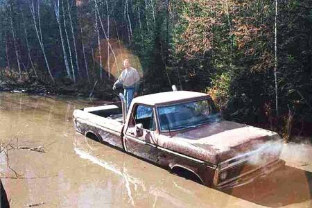 1988 Ford Bronco - A man stands in the bed of a rusted pickup truck that is stuck in a muddy body of water.