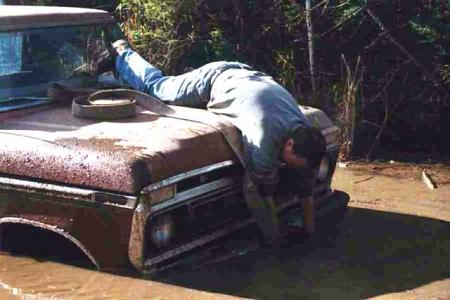 1988 Ford Bronco - Man attempts repairs on a stuck, rusty Ford truck in muddy conditions.