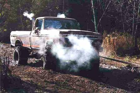 1988 Ford Bronco - A vintage Ford pickup truck navigates a muddy, wooded trail, its exhaust billowing smoke.