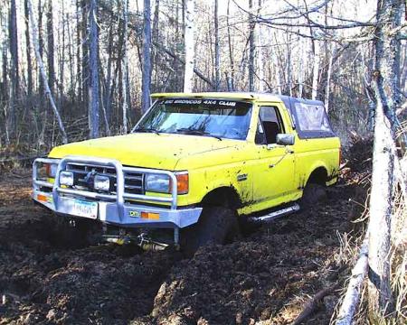 1988 Ford Bronco - A yellow 1988 Ford Bronco is stuck deep in mud amidst a wooded area.
