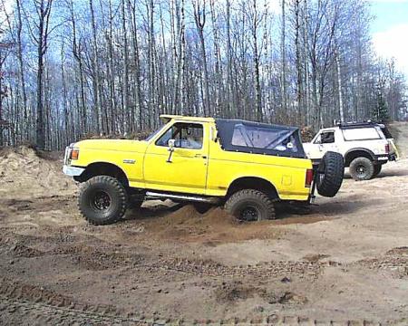 1988 Ford Bronco - Two SUVs are seen off-roading in a wooded area with sandy terrain.