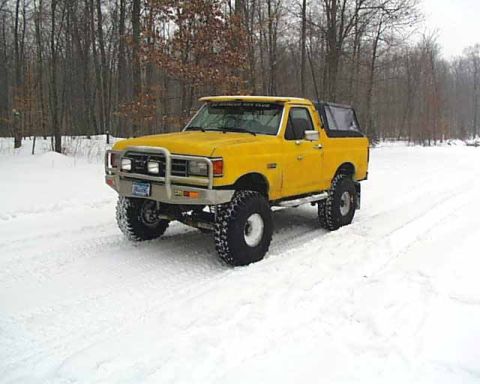 1988 Ford Bronco - A yellow 1987 Ford Bronco with a black soft top and off-road tires drives through a snowy landscape.
