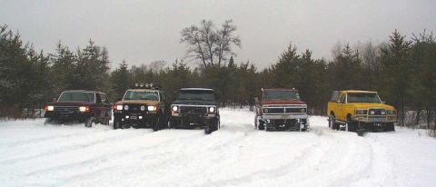1988 Ford Bronco - A group of five SUVs, including a yellow 1988 Ford Bronco, are parked in a snowy landscape with pine trees in the background.
