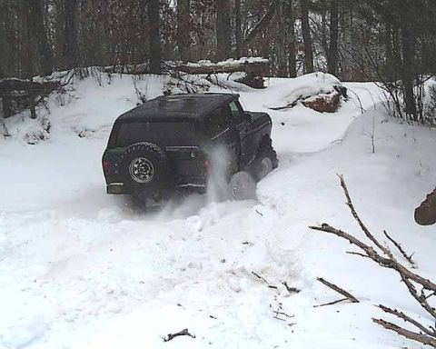 1988 Ford Bronco - A black SUV navigates a snowy, wooded area, with snow spraying from its tires.